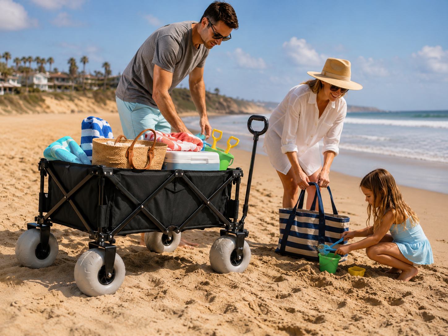 Family preparing beach supplies beside a black foldable beach wagon with large sand wheels on sunny coastal beach, little girl playing sand with toy bucket and shovel