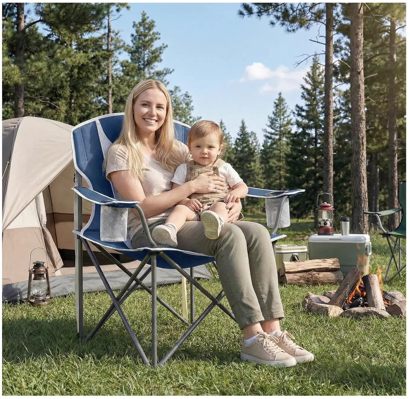Woman sitting in a camping chair holding a baby outdoors with a tent and campfire in the background.
