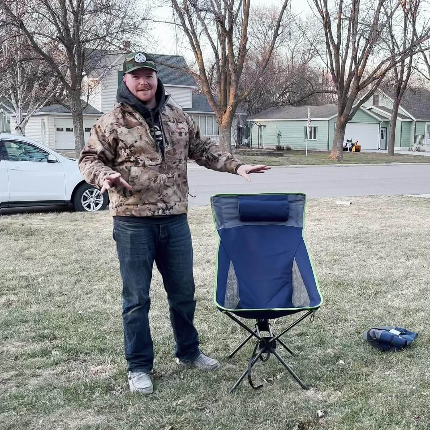 Man in camouflage jacket standing next to a blue folding chair on a grassy area with houses and cars in the background.