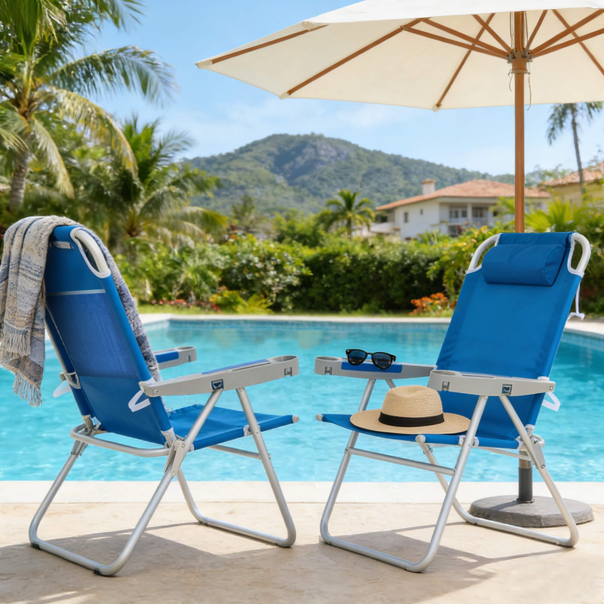 Two blue beach chairs with a sun umbrella by a pool, surrounded by greenery and mountains.
