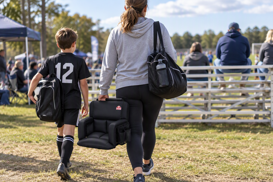 A parent carries a back-support stadium seat and a packed sideline bag toward the bleachers at a youth soccer tournament.