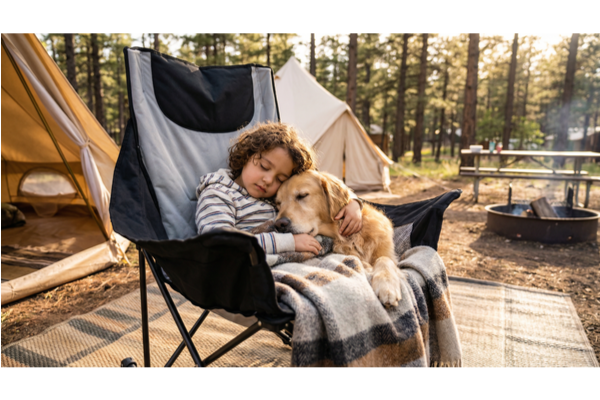 A young child and a golden retriever sleeping together in a high-back padded camping chair at a forest campsite