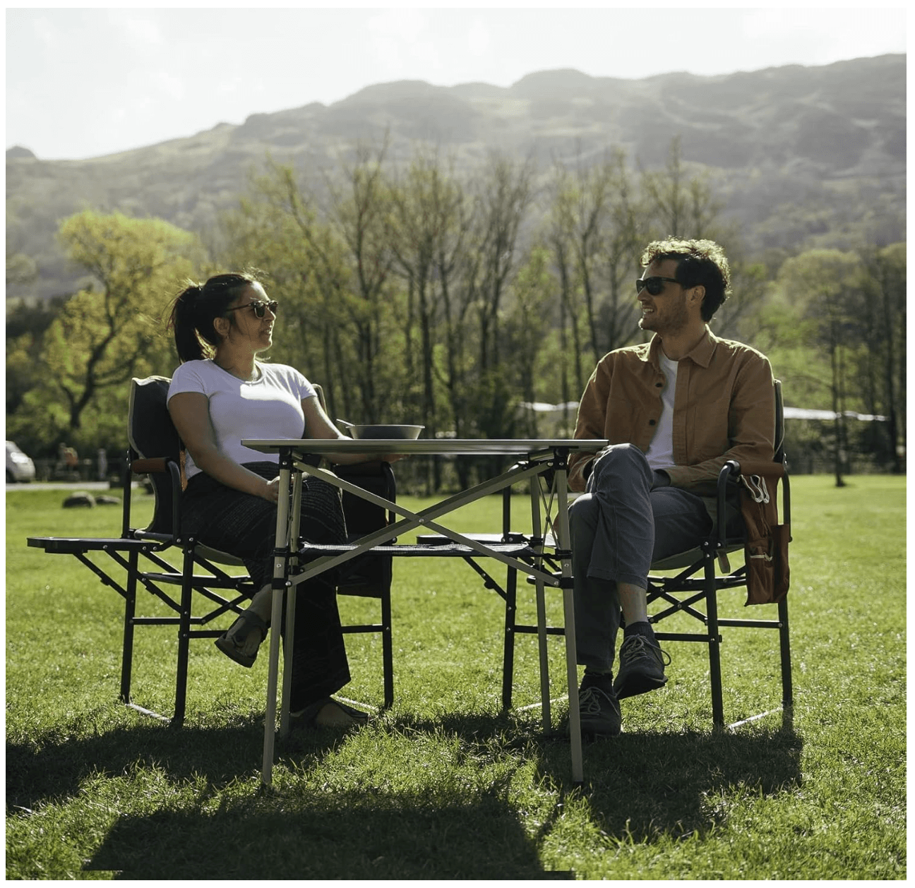 Two people sitting at a picnic table in a park with mountains in the background