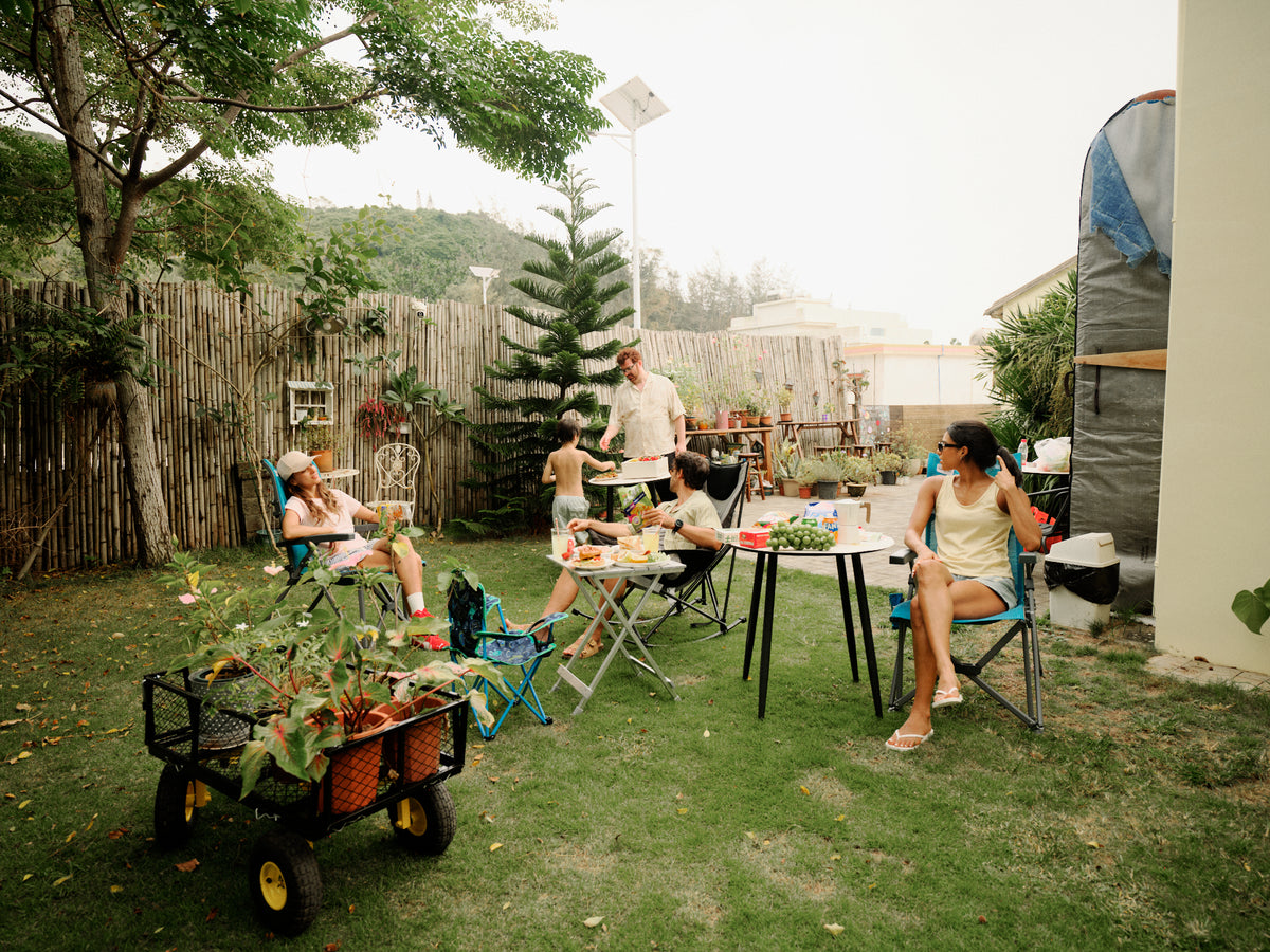 A family gathers in a backyard garden setting, with a woman sitting on a folding chair in the foreground, children and a man standing near a small table, and a cart of potted plants in the grass.