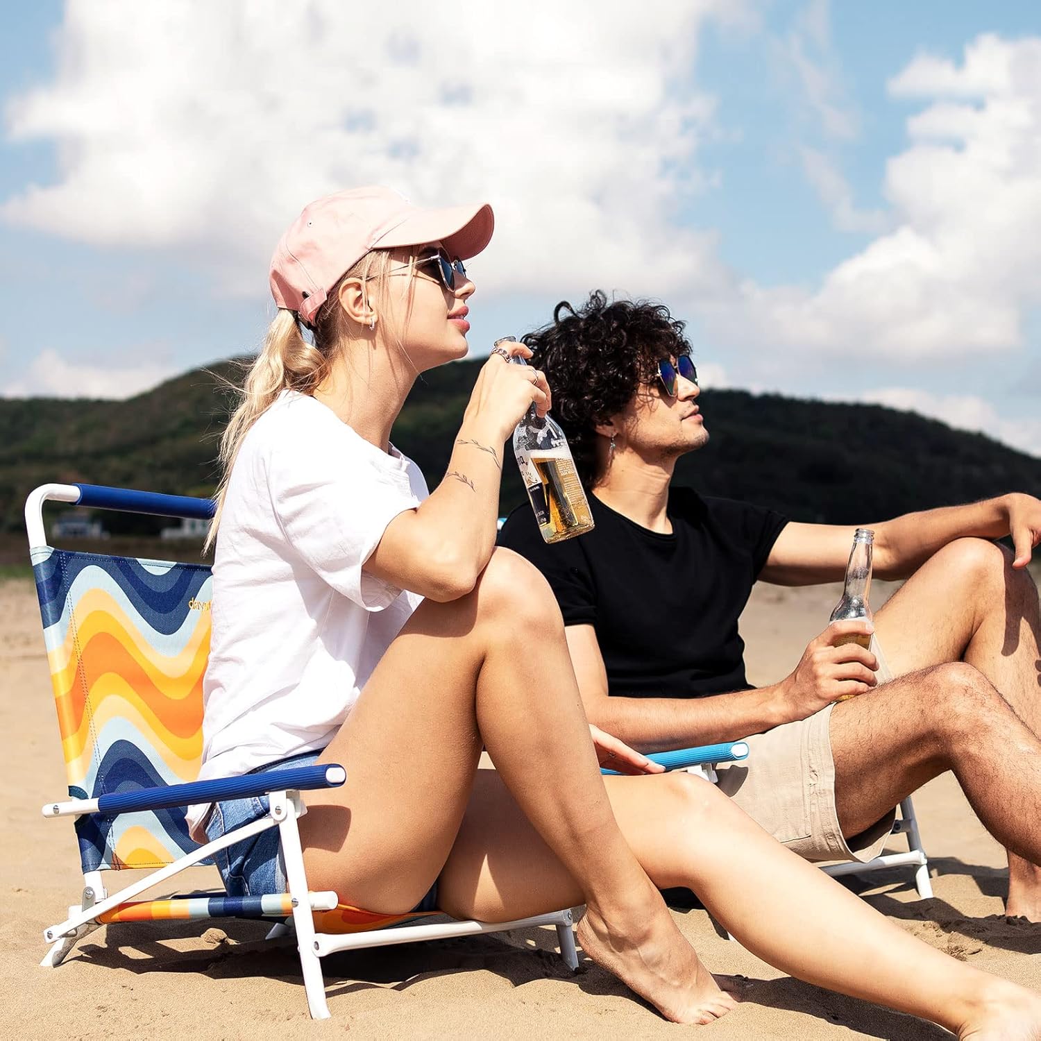 Two people sitting on a beach, one holding a bottle, with a colorful chair and mountainous background.