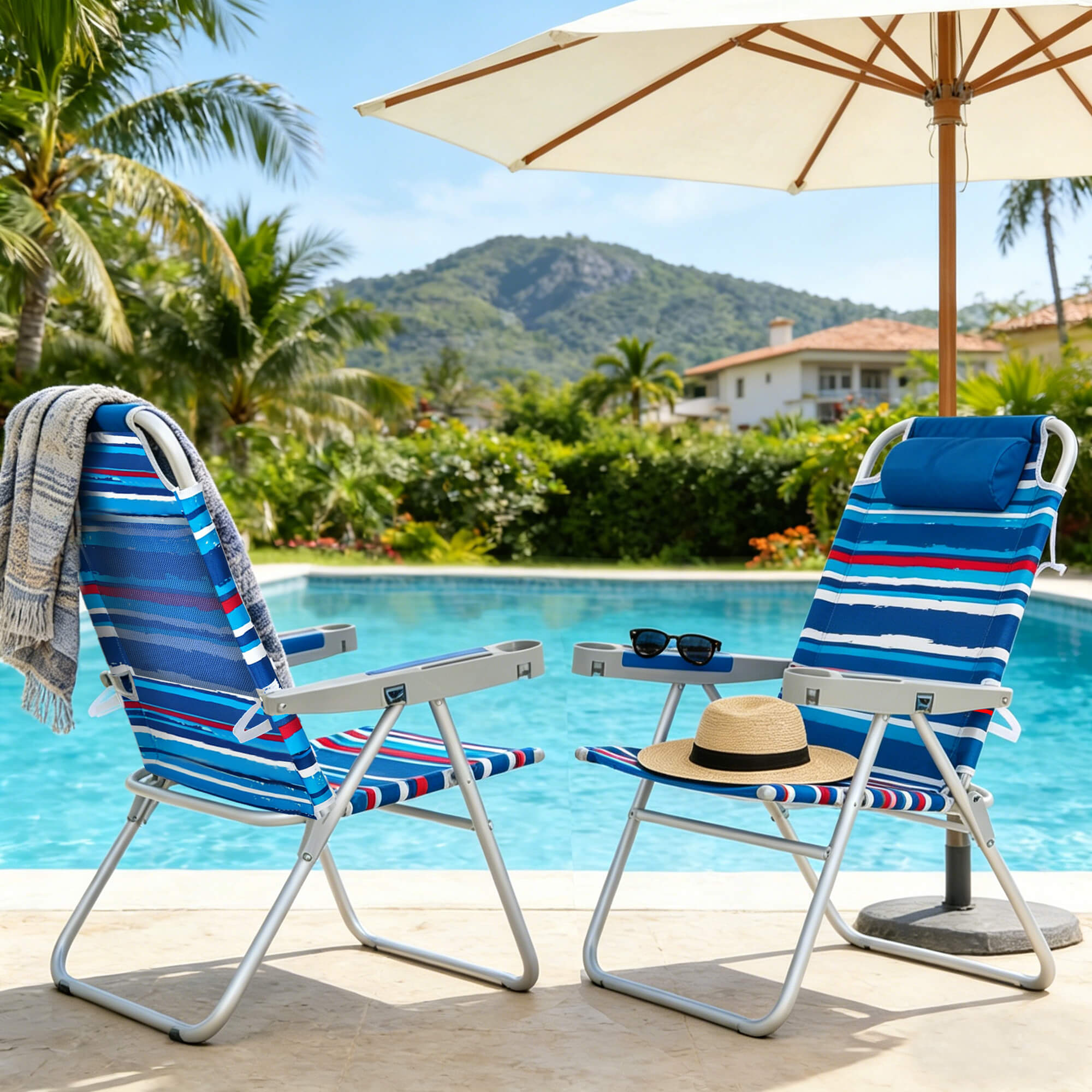 Two blue striped beach chairs with a sun umbrella by a poolside with palm trees and mountains in the background.