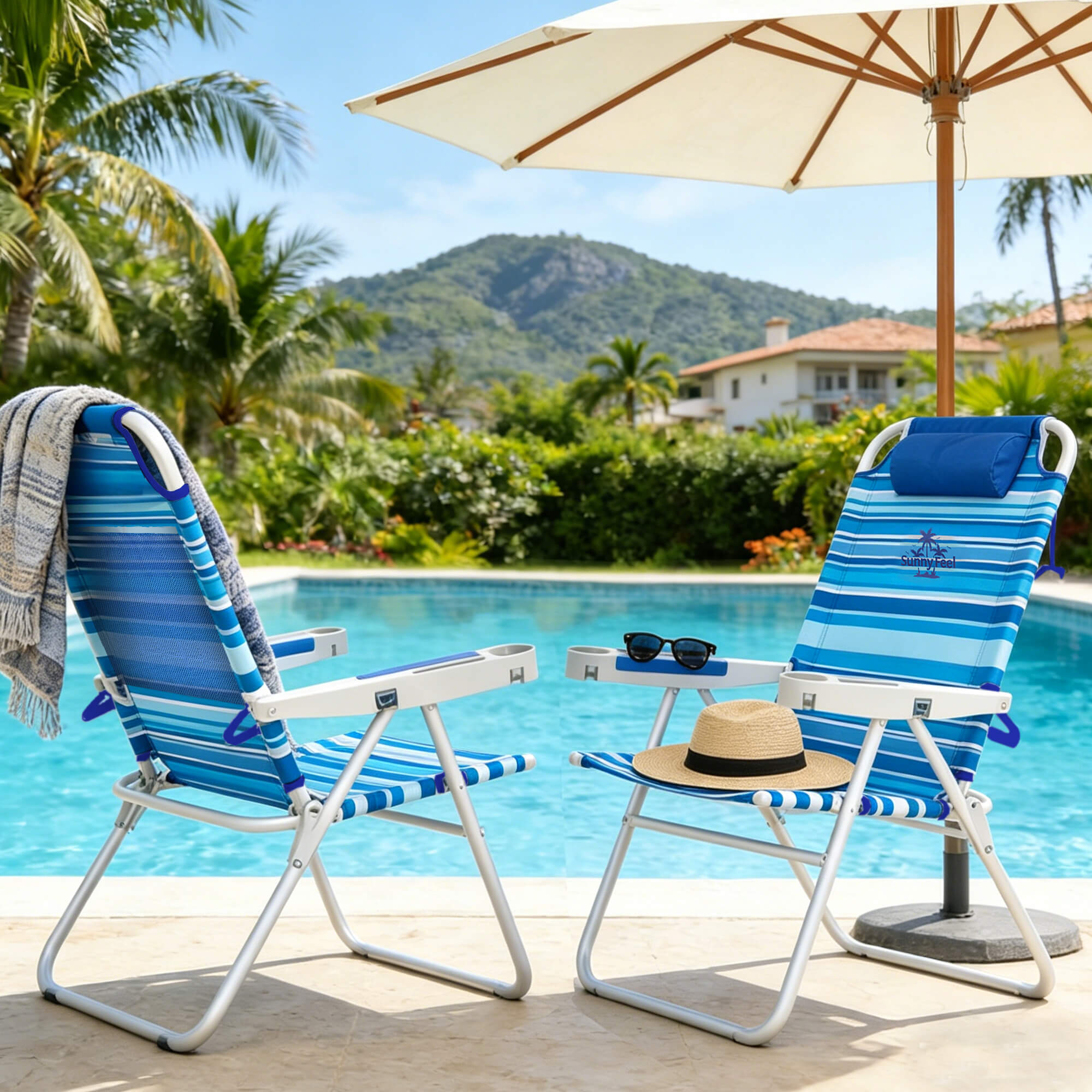 Two blue striped beach chairs with a table and umbrella by a poolside with palm trees and mountains in the background.