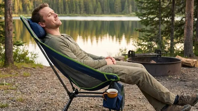 Man relaxing in a camping chair by a lake with a campfire in the background