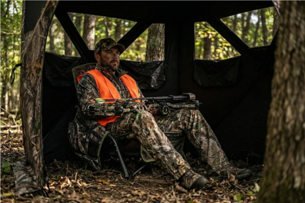 Hunter in camouflage gear and orange vest sitting in a tree stand in the woods.