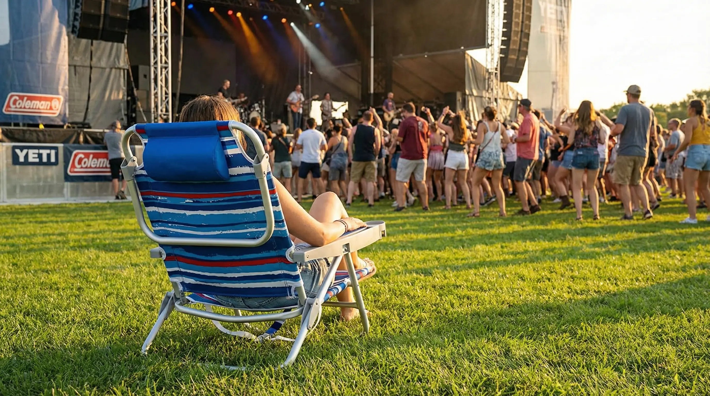 Person sitting in a striped lawn chair watching a concert on a grassy field with stage and people in the background.