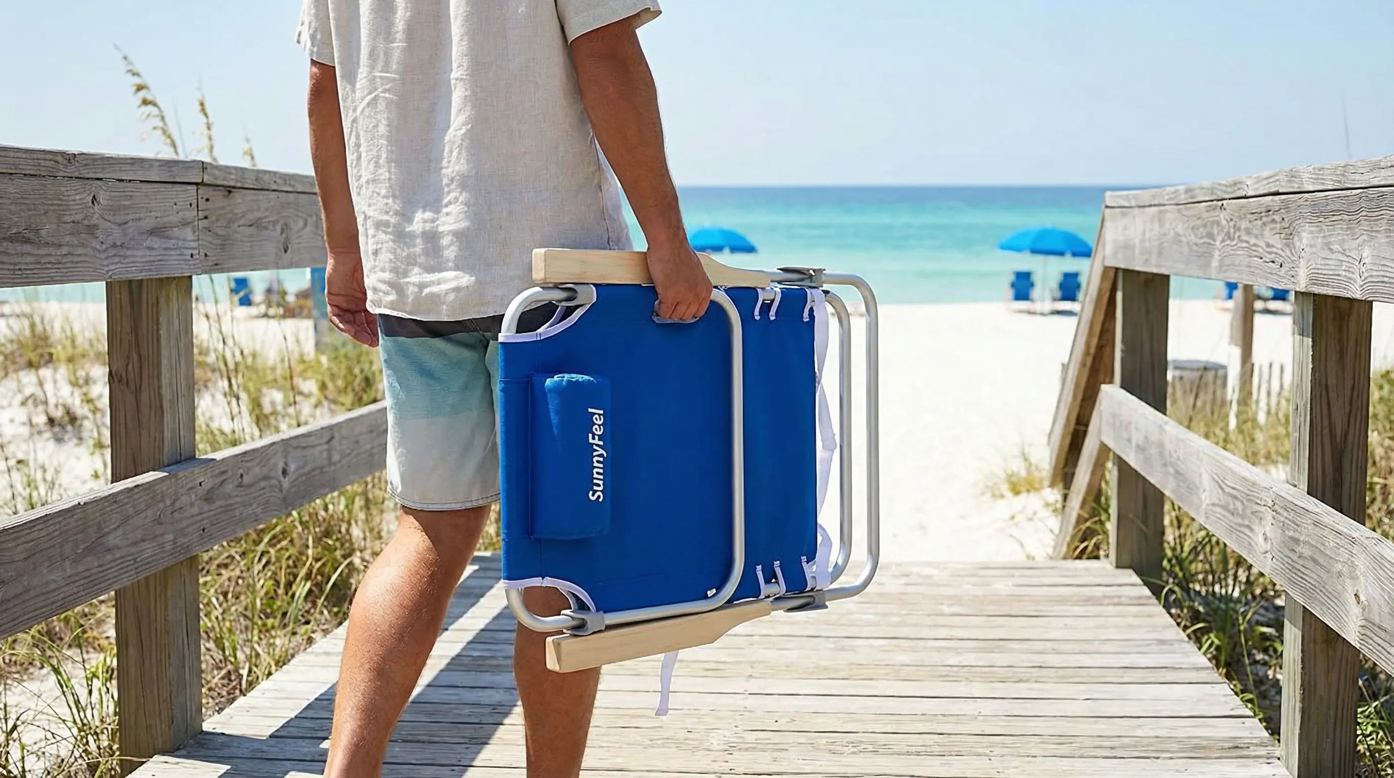 Person holding a blue folding chair on a wooden deck with a beach in the background