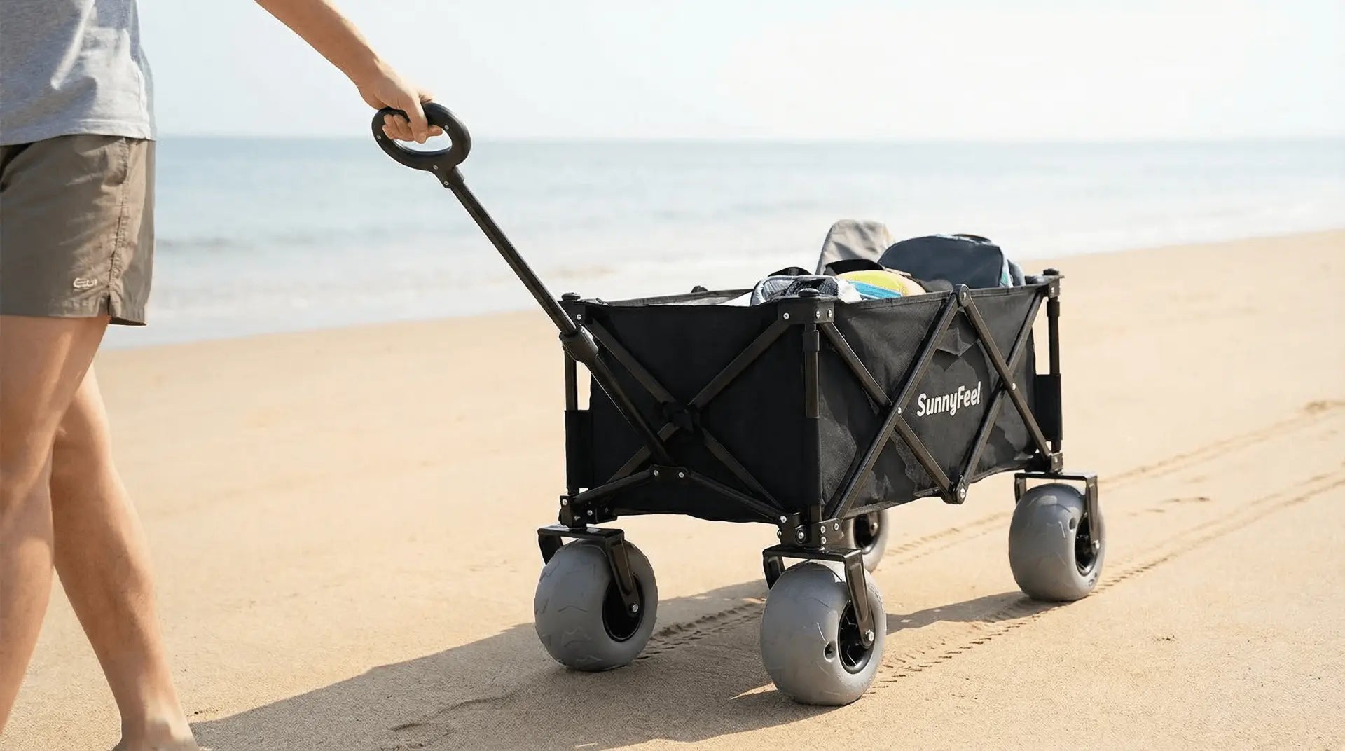 Person pushing a black folding wagon on a sandy beach.